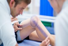 Görselde, bir sağlık uzmanı hastanın bükülen diz ekleminin açısını özel bir ölçüm cihazıyla (gonyometre) dikkatlice ölçüyor. Hasta muayene masasında yatarken ölçüm işlemi yakından yapılıyor.