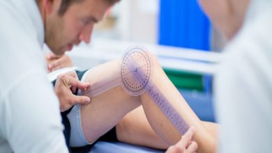 Görselde, bir sağlık uzmanı hastanın bükülen diz ekleminin açısını özel bir ölçüm cihazıyla (gonyometre) dikkatlice ölçüyor. Hasta muayene masasında yatarken ölçüm işlemi yakından yapılıyor.