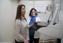 Bir sağlık merkezinde, bir kadın mamografi cihazında tarama için pozisyon alırken, yanında sağlık personeli süreci dikkatle takip ediyor.