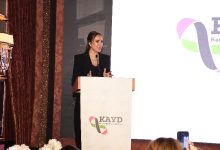 Görselde bir kadın, kürsüde konuşma yaparken görülüyor. Siyah bir takım elbise giymiş, saçları toplu ve mikrofon aracılığıyla dinleyicilere hitap ediyor. Kürsünün üzerinde bir dernek logosu yer alıyor. Arka planda büyük bir ekran ve sahne düzeni bulunuyor. Ön tarafta izleyicilerden bazıları konuşmayı telefonlarıyla kaydediyor.