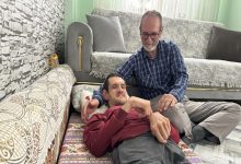 Bir ev ortamında, yerde uzanan genç bir adamın yanında oturan bir kişi ona destek olur gibi omzuna dokunuyor. Arka planda koltuk ve yastıklar yer alıyor. Sahne, aile içi dayanışma ve bakım anını yansıtan sıcak ve samimi bir kare.