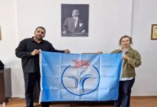 Fotoğrafta bir kadın ve bir erkek, üzerlerinde Yükseköğretim Kurulu (YÖK) logosu bulunan açık mavi bir bayrağı birlikte tutarken görülüyor.