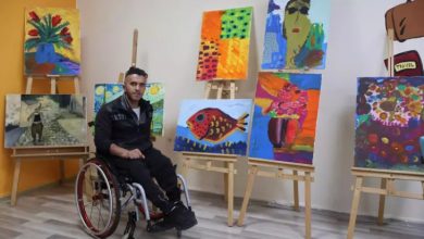 Bedensel engelli bir sanatçının, tekerlekli sandalyesinde oturduğu ve kendi yaptığı rengârenk tablolarla çevrelendiği bir sanat atölyesi karesi. Arka planda şövaleler üzerinde sergilenen balık, çiçek ve portre temalı canlı çalışmalar, atölyenin yaratıcı atmosferini yansıtıyor.