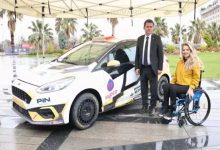 Görselde, engelli ralli pilotu Kübra Denizci Keskin ile Osmangazi Belediye Başkanı Erkan Aydın yan yana görülüyor. Keskin, tekerlekli sandalyesinde otururken kamera karşısına gülümseyerek poz veriyor. Yanlarında, yarışlar için hazırlanmış sponsorluk logoları taşıyan bir ralli aracı bulunuyor. Araç, özel kaplamasıyla profesyonel bir yarış otomobili görünümünde. Arka planda ise palmiyelerin yer aldığı açık alan dikkat çekiyor. Kare, spor alanında erişilebilirliğin ve engelli sporcuların profesyonel arenadaki varlığının önemine işaret eden bir buluşmayı yansıtıyor.