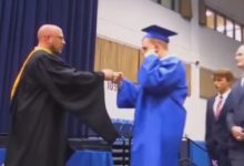 Bir mezuniyet töreni sahnesi: Mavi cübbe ve kep giymiş bir öğrenci, sahnedeki görevliyle yumruk tokuşturuyor. Yan tarafta sırada bekleyen diğer öğrenciler var. Geleneksel tokalaşma yerine daha samimi ve farklı bir kutlama anı yakalanmış.