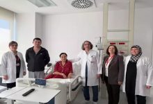 Bir hastane odasında, hasta yatağında oturan bir kadın ve etrafında doktorlar ile sağlık çalışanları birlikte poz veriyor. Ortamda tıbbi cihazlar ve hastane ekipmanları bulunuyor. Sahne, başarılı bir tedavi sonrası çekilmiş, umut ve iyileşme hissi taşıyan bir anı yansıtıyor.