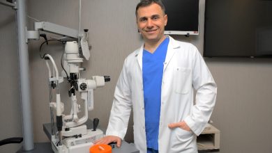 Bir göz muayenehanesinde, beyaz önlüklü bir doktor gelişmiş göz muayene cihazının yanında poz veriyor. Modern tıbbi ekipmanlarla donatılmış ortam, profesyonel ve güven veren bir sağlık hizmeti atmosferi sunuyor.