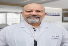 Bir hastane ortamında çekilmiş portre: Beyaz önlük giymiş, orta yaşlı bir doktor kameraya sakin ve güven veren bir ifadeyle bakıyor. Arka planda sağlık kuruluşuna ait bir alan bulanık şekilde görünüyor; profesyonel ve kurumsal bir atmosfer var.