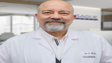 Bir hastane ortamında çekilmiş portre: Beyaz önlük giymiş, orta yaşlı bir doktor kameraya sakin ve güven veren bir ifadeyle bakıyor. Arka planda sağlık kuruluşuna ait bir alan bulanık şekilde görünüyor; profesyonel ve kurumsal bir atmosfer var.