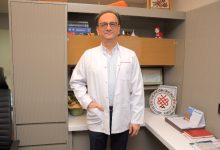 Görselde, bir doktor muayenehane ortamında ayakta duruyor. Beyaz önlük giyen, gözlüklü orta yaş grubunda bir erkek görünüyor. Arkasında kitapların, belgelerin ve dekoratif objelerin bulunduğu raflı bir çalışma alanı yer alıyor. Doktor doğrudan kameraya bakarak sakin bir duruş sergiliyor.