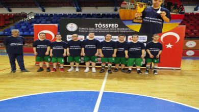 Bir spor salonunda, aynı formayı giymiş bir basketbol takımı saha ortasında yan yana dizilmiş. Arkalarında federasyon logosu ve Türk bayrağı bulunuyor. Görselin köşesinde ise kupayla poz veren bir sporcunun yer aldığı küçük bir ek fotoğraf var. Atmosfer, şampiyonluk gururu ve takım başarısını yansıtıyor.