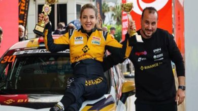 Ralli Pilotu Kübra Denizci Keskin, aracın üstünde oturarak kupaları elinde tutuyor ve yanında erkek bir görevli bulunuyor.