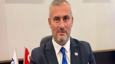 Orta yaşlı bir erkek, resmi bir ortamda masa başında oturuyor. Lacivert takım elbise, beyaz gömlek ve mavi kravat takmış. Saçları kısa ve griye dönmüş, yüzünde hafif sakal var. Arkasında Türk bayrağı ve bir kurum bayrağı görünüyor; genel atmosfer bir ofis ya da resmi kurum ortamını andırıyor.