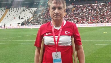 Bir stadyumda, koltuk değnekleriyle saha kenarında duran bir adam, üzerinde Türk bayrağı bulunan milli takım formasıyla poz veriyor. Boynundaki akreditasyon kartı ve arka plandaki tribünler, önemli bir spor organizasyonunun atmosferini yansıtıyor. Sahaya bazen ayakla değil, yürekle çıkılır.