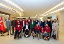 Şık bir salon ortamında, arka planda Türk bayrağının yer aldığı bir mekânda çekilmiş toplu bir fotoğraf. Önde tekerlekli sandalyede oturan sporcular (kırmızı eşofmanlı), arkada ise takım üyeleri ve resmi görünümlü kişiler yan yana poz veriyor. Genel atmosfer resmi ama samimi; bir başarı ya da ziyaret anı gibi duruyor.