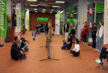 Görselde, bir spor etkinliği alanında oturarak voleybol oynayan kişiler görülüyor. Katılımcılar yerde oturur pozisyonda top alışverişi yaparken çevrede izleyenler ve destek olan kişiler bulunuyor. Etkinlik alanı Intercity logoları ve yeşil bayraklarla süslenmiş, enerjik ve hareketli bir atmosfer yansıtıyor.