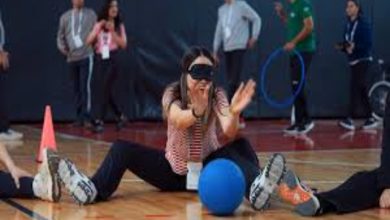 Bir spor salonunda, gözleri siyah bir bantla kapatılmış bir kadının yerde oturarak mavi bir topla paralimpik spor denemesi yaptığı anı gösteren bir fotoğraf. Arka planda etkinliği izleyen ve destek veren diğer katılımcılar ile spor ekipmanları görülüyor. Görsel, engelli sporlarına yönelik bir farkındalık eğitimini veya deneyimleme anını yansıtıyor.