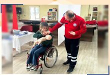 Bir eğitim ortamında, tekerlekli sandalyede oturan bir kadın bebek mankeni üzerinde ilk yardım uygulaması yapıyor. Yanında duran eğitmen ise adım adım nasıl müdahale etmesi gerektiğini gösteriyor. Ortam düzenli ve sakin; masa üzerinde eğitim malzemeleri yer alıyor. Sahne, öğrenme ve pratik yapma anını yansıtıyor.