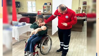 Bir eğitim ortamında, tekerlekli sandalyede oturan bir kadın bebek mankeni üzerinde ilk yardım uygulaması yapıyor. Yanında duran eğitmen ise adım adım nasıl müdahale etmesi gerektiğini gösteriyor. Ortam düzenli ve sakin; masa üzerinde eğitim malzemeleri yer alıyor. Sahne, öğrenme ve pratik yapma anını yansıtıyor.