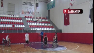 Görselde, bir spor salonunda tekerlekli sandalye basketbol takımı antrenman yapıyor. Oyuncular sahada dağılmış hâlde basket potasına doğru çalışırken, ortada bir antrenör onlara yönlendirme yapıyor. Tribünlerde kırmızı-beyaz koltuklar, yan duvarda Türk bayrağı ve yukarıda “GAZİANTEP” yazılı konum etiketi yer alıyor. Atmosfer, maç öncesi ciddi ve hazırlığa odaklı bir çalışma izlenimi veriyor.