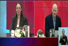 Bir televizyon programına ait ekran görüntüsü. Solda otizmli araştırmacı-yazar Espina Hande Ayas konuk, masada çiçek ve kitaplarla birlikte kameraya dönük konuşuyor. Sağda erkek sunucu Uğur Becerikli elindeki kitaptan okuyor. Alt köşede ise işaret dili tercümanı yer alıyor. Arka plan kırmızı tonlarda stüdyo dekoru.