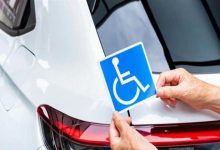 Bir elin, beyaz bir aracın arkasına mavi renkli engelli sembolü (tekerlekli sandalye ikonu) yapıştırdığı veya tuttuğu anı gösteren yakın çekim görsel. Fotoğraf, engelli bireylere yönelik araç kullanım haklarını veya muafiyetlerini temsil eden sembolik bir anlatıma sahip.