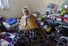 Mütevazı bir ev ortamında yaşlı bir kadın yerde otururken görülüyor. Etrafında günlük yaşam eşyaları, ilaçlar ve yürüme desteği sağlayan ekipmanlar bulunuyor. Görsel, sağlık sorunları nedeniyle zor koşullarda sürdürülen yaşam mücadelesini yansıtıyor.