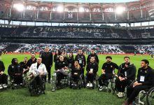 Görselde, bir futbol stadyumunun çim sahasında bir araya gelmiş büyük bir grup görülüyor. Ön sırada tekerlekli sandalyede bulunan sporcular yer alırken, arkalarında teknik ekip ve yöneticiler ayakta duruyor. Sporcuların çoğunun elinde kupa ve çiçek buketleri bulunuyor. Arka planda tribünlerde “Tüpraş Stadyumu” yazısı seçiliyor. Atmosfer, bir kutlama ve onurlandırma töreni havası taşıyor.