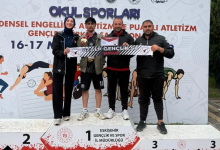 Bir spor müsabakası ödül töreni görülüyor. Üç basamaklı bir kürsü üzerinde dört kişi ayakta duruyor. Ortadaki sporcu birincilik basamağında ve boynunda madalya taşıyor. Grup, “ÖZALP GENÇLİK SK ŞAHİNLER” yazılı bir pankart tutuyor. Arkadaki afişte “Okul Sporları”, “Bedensel Engelliler Atletizm ve Puanlı Atletizm Gençlik Spor Şampiyonası” ifadeleri ve “16–17 Mayıs” tarihi yer alıyor. Arka planda sporcu illüstrasyonları bulunuyor. Atmosfer başarı ve gurur duygusu taşıyor.