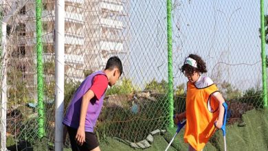 Fotoğrafta, iki çocuk açık havada bir futbol sahasında birlikte top oynarken görülüyor. Çocuklardan biri ampute ve koltuk değnekleriyle hareket ediyor; üzerinde turuncu bir yelek var. Diğer çocuk ise mor yelek giyiyor ve topa doğru yönelmiş durumda. Arka planda tel örgüler ve yüksek bir bina dikkat çekiyor.