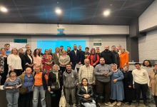 Bir konferans salonunda, farklı yaş gruplarından kalabalık bir grup sahne önünde toplu fotoğraf veriyor. Arka planda sunum ekranı yer alırken, katılımcılar yan yana dizilmiş. Aralarında tekerlekli sandalye kullanan bireyler de bulunuyor. Ortam, bir zirve ya da eğitim etkinliği sonrası çekilen hatıra fotoğrafı havası taşıyor.