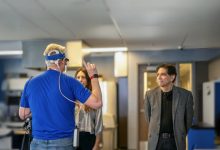 Kapalı bir laboratuvar ortamında başına elektronik cihaz takılı bir kişi el hareketiyle bir şey anlatırken, karşısında duran iki kişi dikkatle onu izliyor. Teknolojik ekipmanların bulunduğu sahne, bilimsel bir test ya da araştırma çalışmasını yansıtıyor.