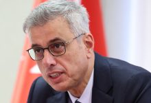 Bakan Memişoğlu konuşma yaparken görülüyor. Arka planda kırmızı‑beyaz bir bayrak bulunuyor; sahne resmi ve ciddi bir ortam hissi veriyor.