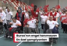 Kapalı spor salonunda düzenlenen etkinlikte tekerlekli sandalye kullanan bireyler ve gönüllüler Türk bayraklarıyla yürüyüş yapıyor. Katılımcıların büyük bölümü beyaz tişört ve kırmızı yelekler giyerken, etkinlik coşkulu ve dayanışma dolu bir atmosfer yansıtıyor.