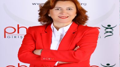 Görselde, kırmızı bir takım elbise ve beyaz gömlek giyen bir kadın kamera karşısında poz veriyor. Kollarını çapraz şekilde bağlamış, gülümseyen bir ifadeye sahip. Arka planda bir derneğe ait olduğu anlaşılan logo ve yazılar bulunuyor.