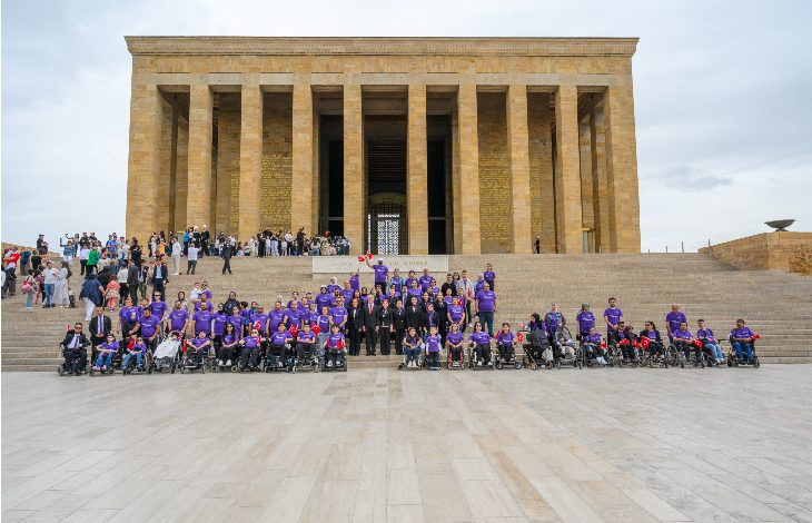 Anıtkabir’in önünde bir araya gelen SMA’lı bireyler, aileleri ve destekçileri toplu fotoğraf çektiriyor. Katılımcıların çoğu mor renkli kıyafetler giyerken, bazı bireyler tekerlekli sandalye kullanıyor. Görsel, birlik, dayanışma ve toplumsal farkındalık mesajı taşıyor.