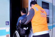 Turuncu yelekli bir görevli, tekerlekli sandalye kullanan bir yolcunun trene binişine yardımcı oluyor. Tren kapısının yanında engelli erişim işareti yer alırken, görüntü erişilebilir ulaşım hizmetlerini yansıtıyor.