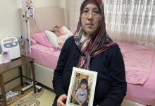 Bir ev ortamında çekilen fotoğrafta, başörtülü bir kadın elinde çerçeveli bir çocuk fotoğrafı tutarak kameraya bakıyor. Arka planda ise tıbbi cihazların bulunduğu bir yatakta yatan bir çocuk görülüyor. Görsel, sağlık sorunlarıyla mücadele eden bir çocuğun ve ailesinin yaşadığı zorlu sürece dikkat çekiyor.