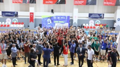 Bu görselde, bir spor salonunda düzenlenen kalabalık bir etkinlik sırasında katılımcılar ellerini havaya kaldırarak coşkulu bir şekilde kutlama yaparken, salonun ortasında bulunan görevliler ve sporcular etkinliğe eşlik etmektedir.