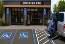Bir marketin önünde yer alan erişilebilir otopark alanları görülüyor. Mavi tekerlekli sandalye sembolleriyle işaretlenmiş park yerlerinin yanında, rampalı bir minibüsten tekerlekli sandalye kullanan bir kadın iniyor. Görsel, erişilebilir park alanlarının ve araç erişim rampalarının önemini vurguluyor.