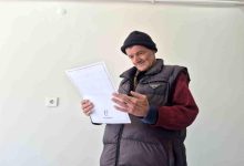 Görselde, bere ve şişme yelek giyen bir kişi elindeki belgeyi dikkatle incelerken görülüyor. Sade ve açık renkli bir odada çekilen fotoğrafta kişinin yüzünde memnuniyet ve rahatlama ifadesi dikkat çekiyor. Belgeye odaklanan kişi ayakta dururken, görüntü önemli bir gelişme veya sevindirici bir haber aldığı anı yansıtıyor.