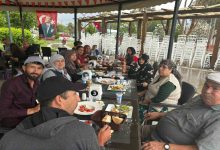 Görselde, açık alanda düzenlenen bir etkinlikte uzun bir masa etrafında bir araya gelen aileler yemek eşliğinde vakit geçiriyor. Türk bayrakları ve doğayla iç içe ortam dikkat çekerken, katılımcıların birlikte zaman geçirdiği samimi ve sosyal bir buluşma atmosferi öne çıkıyor.