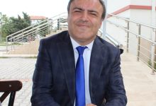 Takım elbiseli bir kişi, dış mekânda merdiven kenarında oturuyor; arka planda açık renk bina ve yeşillik görünüyor.