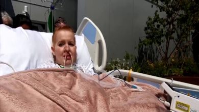 Fotoğrafta hastane yatağında yatan bir hasta görülüyor. Hastanın burnunda ve ağzında tıbbi tüpler bulunuyor; çeşitli kablolar ve monitör bağlantılarıyla tedavisi sürdürülüyor. Üzerine açık pembe renkli bir battaniye örtülmüş durumda. Yatak, kapalı bir yoğun bakım odasında değil, açık havadaki bir bahçe veya teras alanında konumlandırılmış görünüyor. Arka planda bitkiler, çalılar ve dış mekân unsurları dikkat çekiyor. Hastanın yanında sağlık ekipmanları ve serum askısı bulunuyor.
