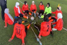 Fotoğrafta ampute kadın futbolcuların sahada daire şeklinde oturarak antrenman veya takım buluşması yaptığı görülüyor. Sporcular turuncu formalar giyiyor ve ortada bir futbol topu bulunuyor. Bazı sporcular protez veya destek ekipmanlarıyla yer alırken, görüntü takım ruhu, dayanışma ve spora katılım temasını öne çıkarıyor.