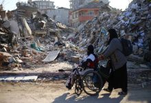 Görselde, tekerlekli sandalyedeki bir kişi ve ona eşlik eden bir kadın, yıkılmış binaların enkazları arasında ilerliyor. Fotoğraf, afetlerin engelli bireyler üzerindeki etkisini ve erişilebilir afet yönetiminin önemini vurgulayan çarpıcı bir sahne sunuyor.