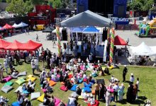 Açık havada düzenlenen etkinlikte insanlar renkli minderlerin üzerinde oturarak sahnedeki programı izlerken, çevrede kurulu çadırlar ve kalabalık bir festival atmosferi dikkat çekmektedir.