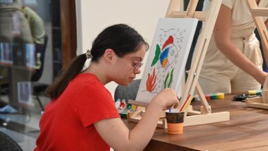 Kırmızı tişörtlü ve gözlüklü bir kız çocuğu, şövale üzerindeki renkli deniz temalı tuvali dikkatle boyarken, arka planda başka bir kişinin de resim yaptığı bir sanat atölyesi ortamı görülmektedir.