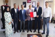 Bir ofis ortamında altı kişi birlikte poz veriyor. Ortadaki iki kişi kırmızı dosya içinde bir belge tutuyor; arka planda Türk bayrağı ve duvarda çerçeveli portreler görülüyor. Görüntü resmi bir belge teslimi ya da açılış/izin töreni izlenimi veriyor.