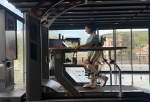 Bir kadın, robotik dış iskelet (ekzoskeleton) desteğiyle bir yürüyüş rehabilitasyon cihazında adım atarken, modern ve geniş pencereli bir ortamda fiziksel terapi çalışması yapıyor.